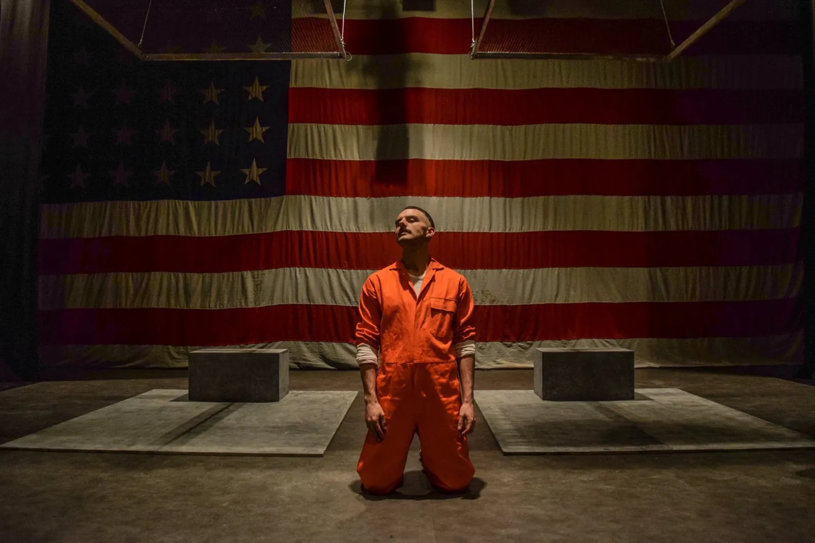 Danny Solomon on stage — orange jumpsuit kneeling, flag backdrop