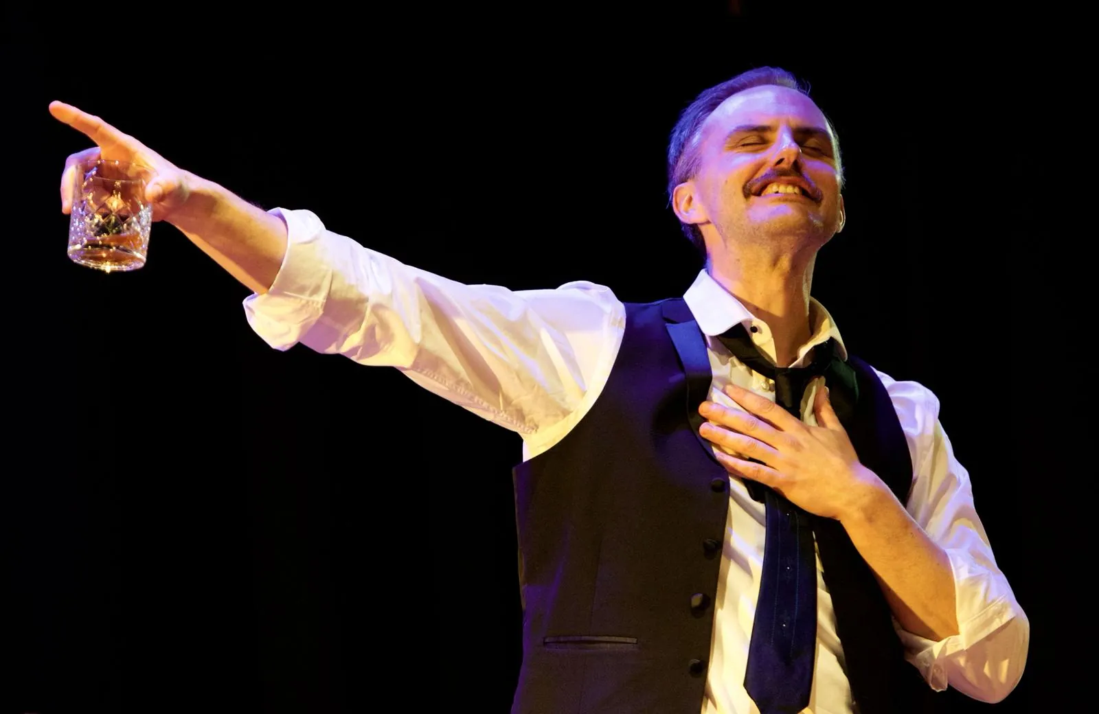 Danny Solomon on stage — pointing with a glass, chandelier-lit room