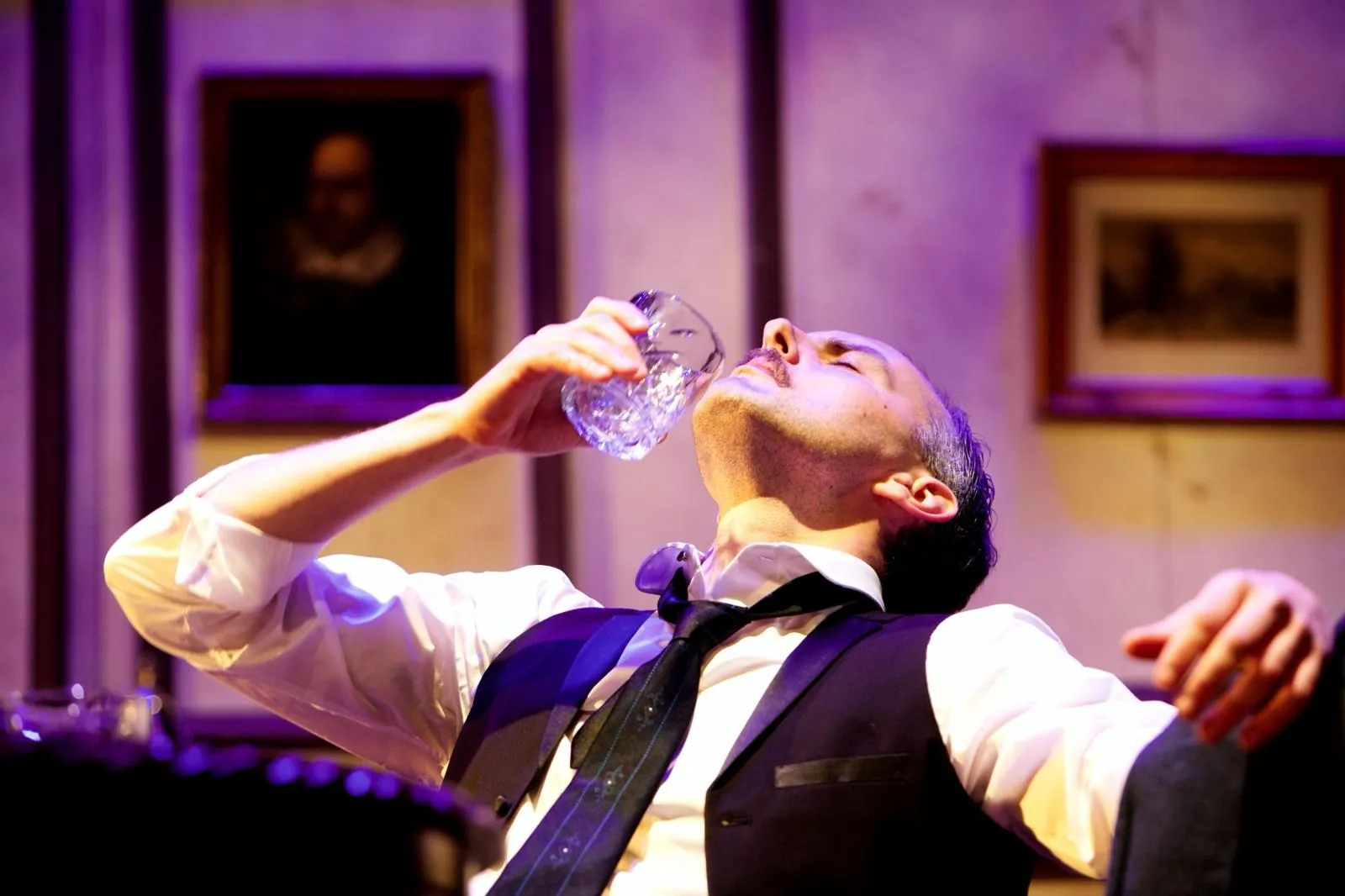 Danny Solomon on stage — drinking, portrait-lined wall