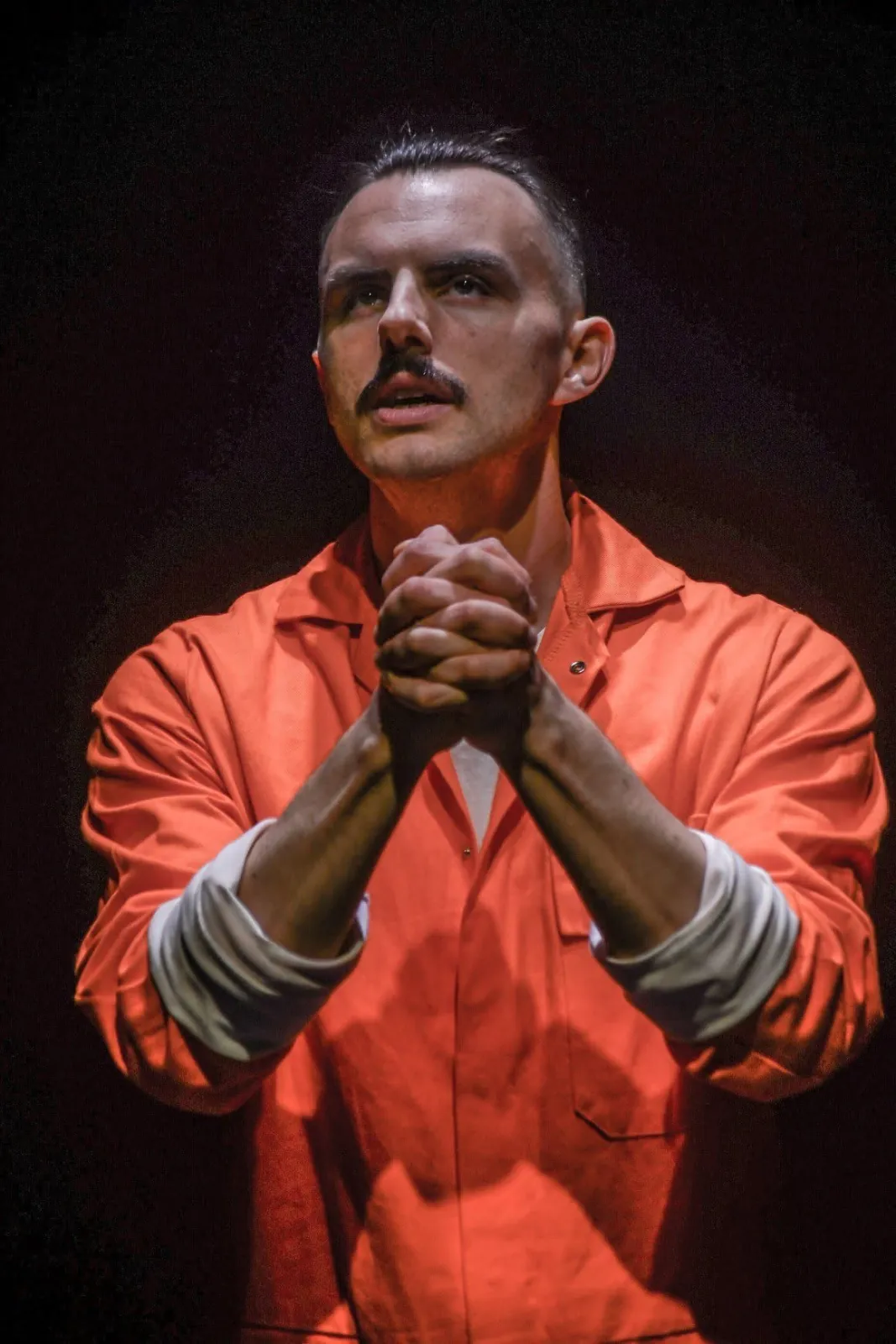 Danny Solomon on stage — orange jumpsuit, hands clasped, spotlit
